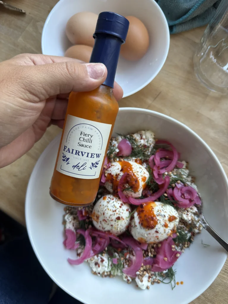 Katinka hält eine Flasche Fiery Chilli Sauce vom Weingut Fairview über den Teller mit weichen Eiern