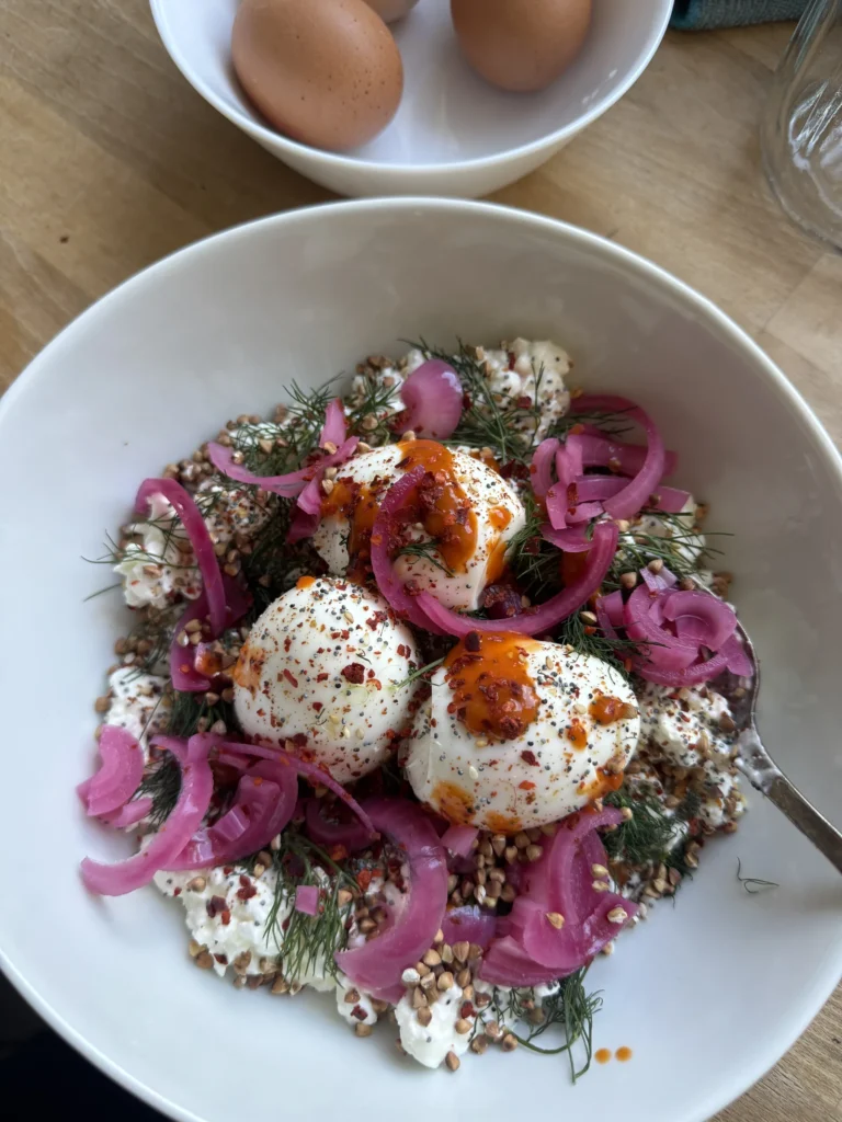 Weiße Schale von oben mit Hüttenkäse-Feta-Creme, drei weichen Eiern, pink eingelegten Zwiebeln, Dill, geröstetem Buchweizen und Chilisauce